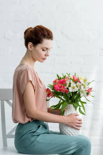 Vue latérale d'une jolie jeune femme tenant un vase avec des fleurs tout en étant assis sur une chaise devant un mur de briques blanches — Photo de stock