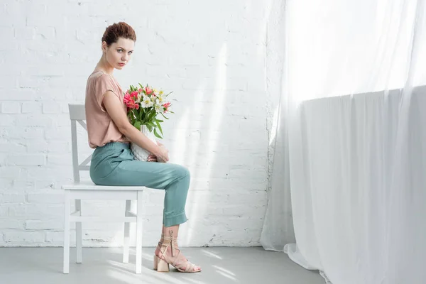 Sensual young woman with flowers in vase in front of white brick wall — Stock Photo