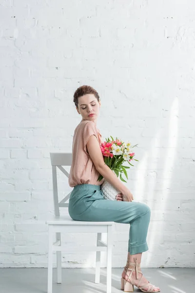 Beautiful young woman with fresh flowers in vase in front of white brick wall — Stock Photo