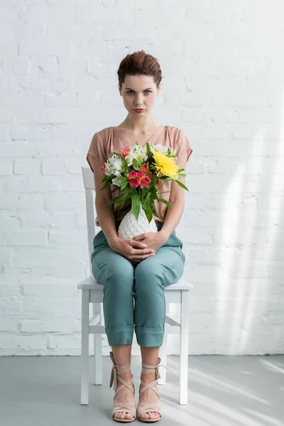 Belle jeune femme avec des fleurs dans un vase devant un mur de briques blanches — Photo de stock