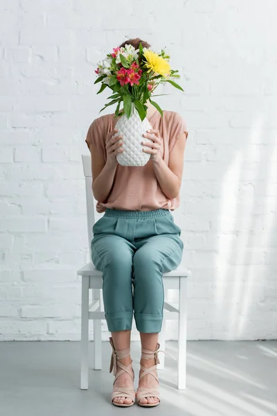 Femme couvrant le visage de fleurs dans un vase assis sur une chaise devant un mur de briques blanches — Photo de stock