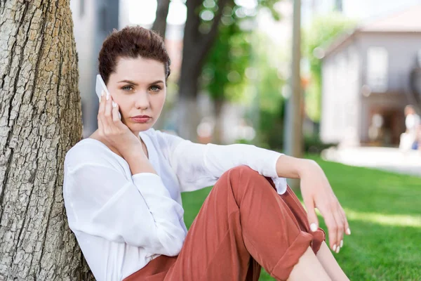 Beautiful young woman talking by phone while leaning back on tree trunk in park and looking at camera — Stock Photo