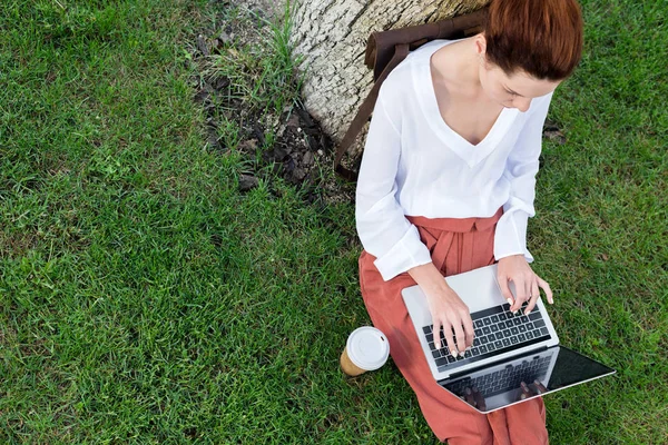 Верхний вид привлекательной молодой женщины, работающей с ноутбуком, сидя на траве с бумажной чашкой кофе — стоковое фото