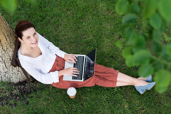 Vista superior de la hermosa mujer joven que trabaja con el ordenador portátil mientras está sentado en la hierba con una taza de papel de café - foto de stock