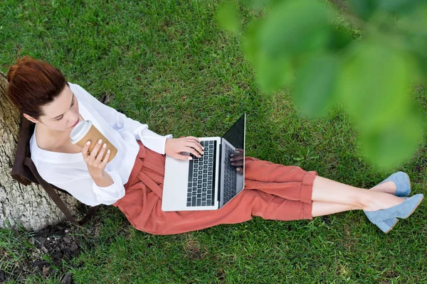 Верхний вид счастливой молодой женщины, работающей с ноутбуком, сидя на траве с бумажной чашкой кофе — стоковое фото