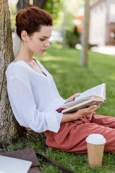 Vista lateral da jovem mulher inclinando-se para trás no tronco da árvore no parque e livro de leitura — Fotografia de Stock