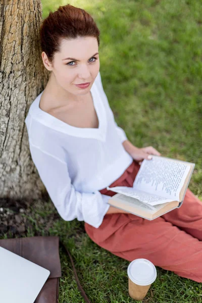 Vista de ángulo alto de la joven feliz con libro apoyado en el tronco del árbol en el parque y mirando a la cámara - foto de stock