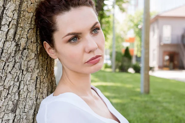 Close-up shot of thoughtful young woman leaning back on tree trunk in park and looking away — Stock Photo