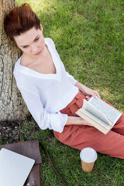Vista de ángulo alto de la hermosa mujer joven con libro apoyado de nuevo en el tronco del árbol en el parque - foto de stock