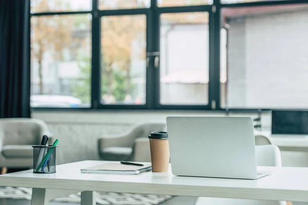 Portatile, tazza usa e getta, notebook e portapenne su tavolo bianco in ufficio moderno — Foto stock