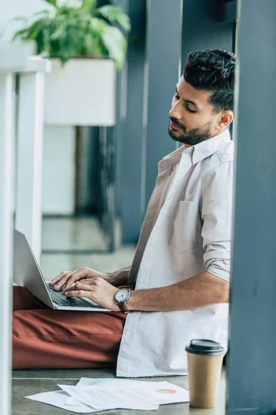 Hombre de negocios reflexivo en ropa casual usando el ordenador portátil mientras está sentado en el suelo en la oficina - foto de stock