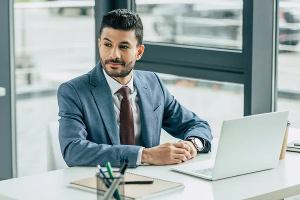 Lächelnder Geschäftsmann schaut weg, während er am Arbeitsplatz in der Nähe von Laptop sitzt — Stockfoto