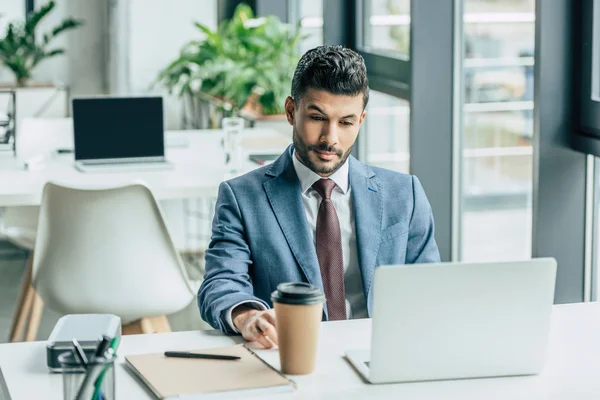 Pensativo hombre de negocios sentado en el lugar de trabajo cerca de portátil y café para ir - foto de stock