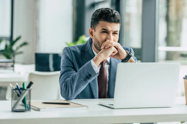 Вдумливий бізнесмен сидить на робочому місці біля ноутбука і тримає складені руки біля обличчя — стокове фото
