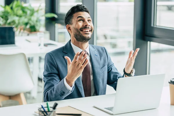 Homme d'affaires souriant montrant s'il vous plaît geste et levant les yeux tout en étant assis sur le lieu de travail — Photo de stock