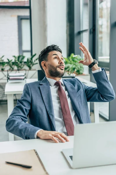 Hombre de negocios emocionado mostrando gesto de idea mientras está sentado en el lugar de trabajo y mirando hacia arriba - foto de stock