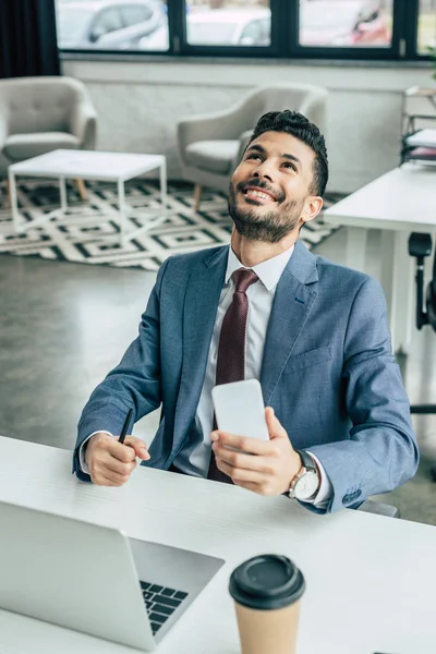 Счастливый бизнесмен смотрит вверх, сидя рядом с ноутбуком и держа в руках смартфон — стоковое фото