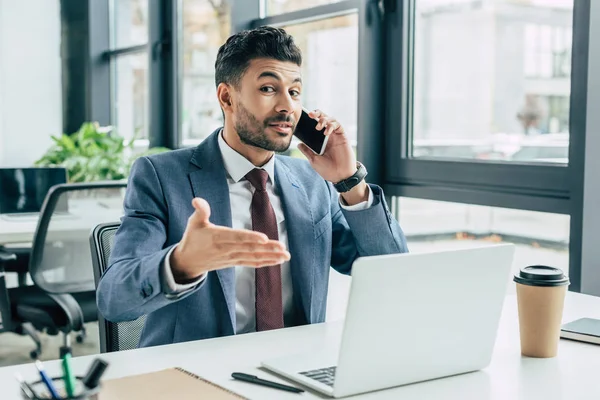 Positive businessman pointing with hand at laptop while talking on smartphone — Stock Photo