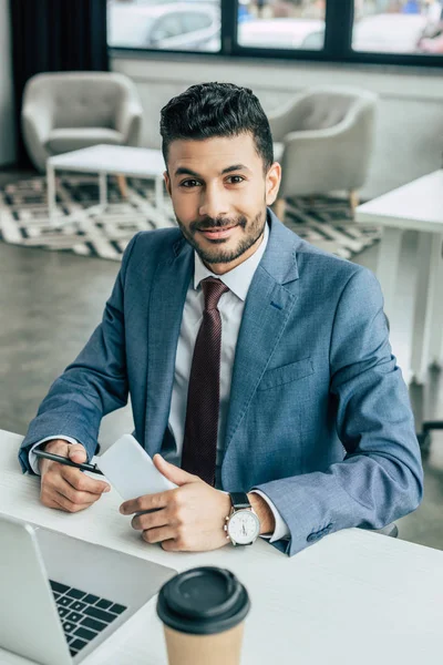 Sonriente hombre de negocios mirando a la cámara mientras está sentado en el lugar de trabajo cerca de la computadora portátil y sosteniendo el teléfono inteligente - foto de stock