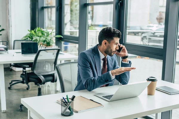 Hombre de negocios concentrado gesto mientras está sentado en el lugar de trabajo y hablando en el teléfono inteligente - foto de stock