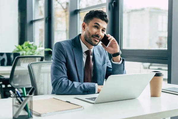 Homme d'affaires souriant parlant sur smartphone tout en étant assis sur le lieu de travail près d'un ordinateur portable — Photo de stock