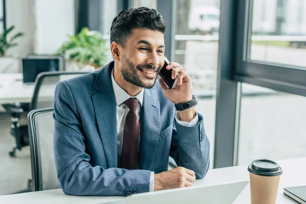 Cheerful businessman talking on smartphone while sitting at workplace in office — Stock Photo