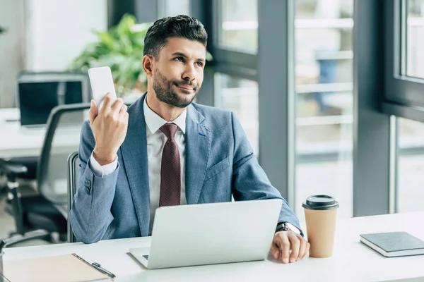 Hombre de negocios aburrido mirando hacia otro lado mientras está sentado cerca de la computadora portátil y sosteniendo el teléfono inteligente - foto de stock