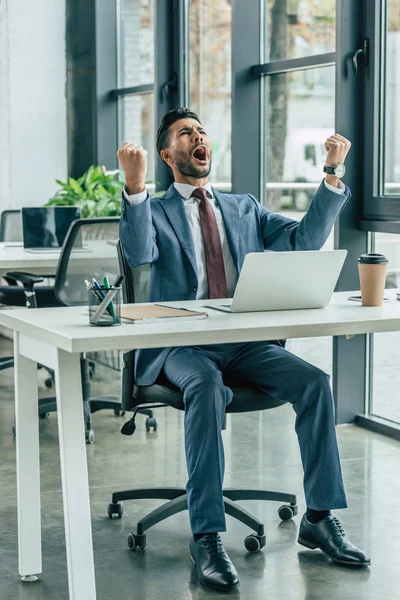 Homme d'affaires heureux crier et montrer geste gagnant tout en étant assis sur le lieu de travail — Photo de stock