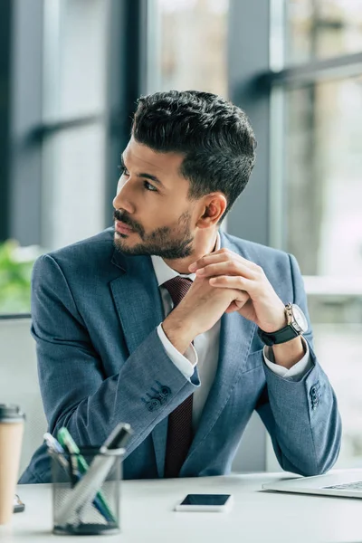 Pensativo hombre de negocios mirando hacia otro lado mientras está sentado en el lugar de trabajo con las manos dobladas - foto de stock