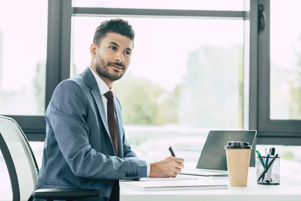 Homme d'affaires souriant écrivant dans un carnet et regardant ailleurs tout en étant assis sur le lieu de travail — Photo de stock