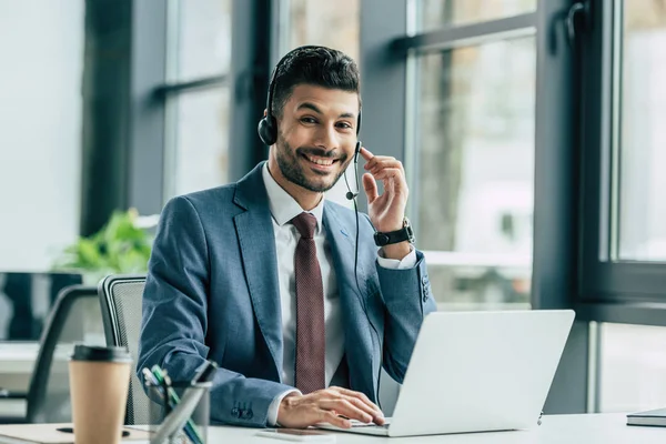 Operatore allegro call center guardando la fotocamera mentre seduto sul posto di lavoro vicino al computer portatile — Foto stock