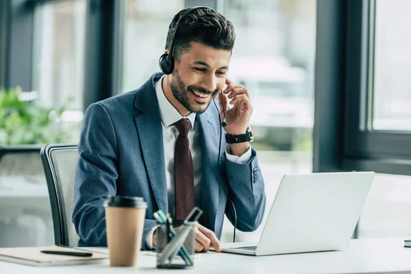 Cheerful call center operator looking at laptop while sitting at workplace — Stock Photo