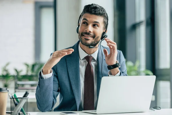 Operador de centro de llamadas sonriente en auriculares mirando hacia arriba mientras está sentado en el lugar de trabajo - foto de stock