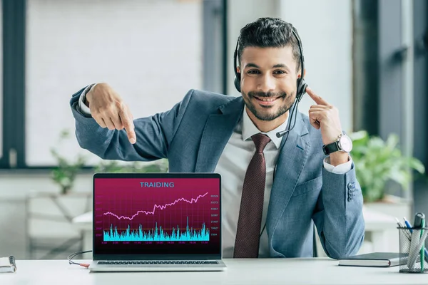 Operador de centro de llamadas alegre apuntando con el dedo a la computadora portátil con infografías comerciales en la pantalla - foto de stock