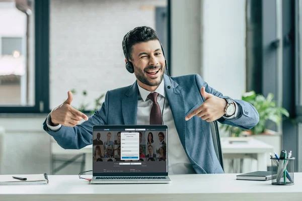 KYIV, UCRANIA - 10 de octubre de 2019: Operador de centro de llamadas alegre guiñando un ojo a la cámara y apuntando con los dedos a la computadora portátil con el sitio web Linkedin en la pantalla . - foto de stock