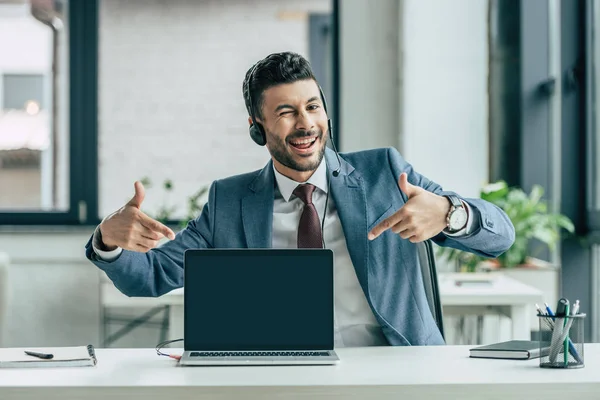 Cheerful call center operator winking at camera and pointing with fingers at laptop with blank screen — Stock Photo