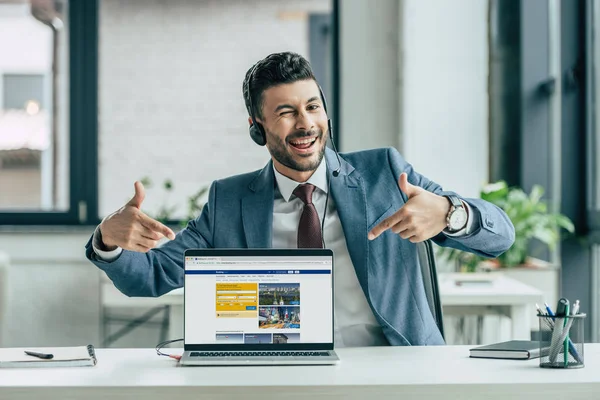 KYIV, UKRAINE - 10 de outubro de 2019: operador de call center alegre piscando para a câmera e apontando com os dedos para o laptop com o site Booking.com na tela . — Fotografia de Stock