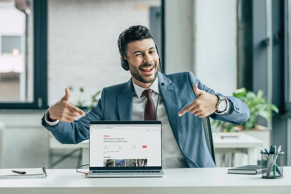 KYIV, UCRANIA - 10 DE OCTUBRE DE 2019: Operador de centro de llamadas alegre guiñando un ojo a la cámara y apuntando con los dedos a la computadora portátil con Airbnb en el sitio web en la pantalla . - foto de stock