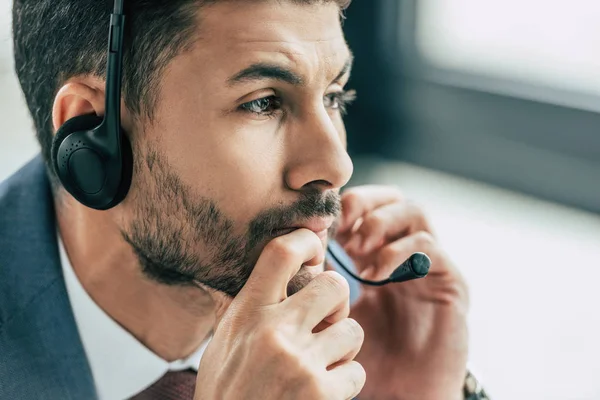 Operador de centro de llamadas reflexivo en auriculares mirando hacia otro lado y sosteniendo la cabeza cerca de la cara - foto de stock