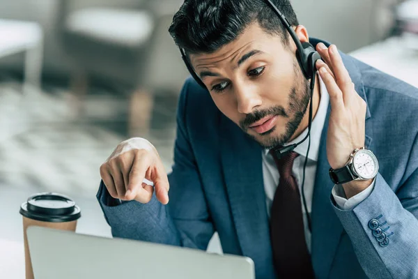 Operador de centro de llamadas concentrado en auriculares apuntando con el dedo a la computadora portátil - foto de stock