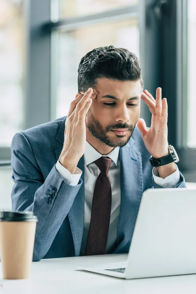 Hombre de negocios agotado agarrado de la mano cerca de la cabeza mientras está sentado en el lugar de trabajo y sufre de migraña - foto de stock