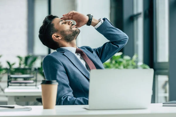 Tired businessman holding hand near head and looking up while sitting at workplace — Stock Photo