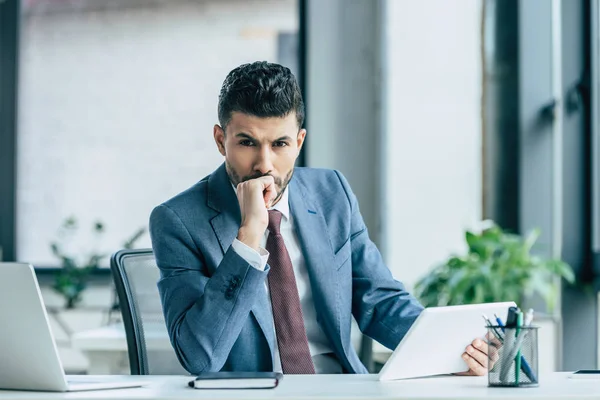 Thoughtful businessman holding laptop and looking at camera while sitting at workplace — Stock Photo