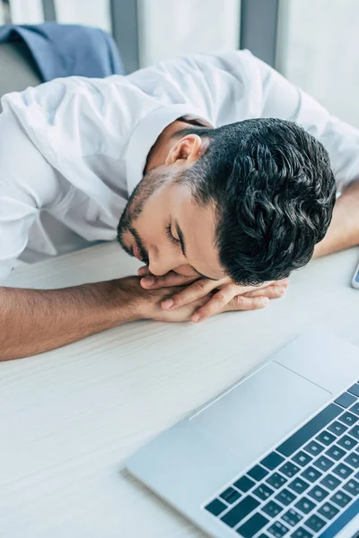 Joven, hombre de negocios agotado durmiendo en el lugar de trabajo cerca de la computadora portátil - foto de stock