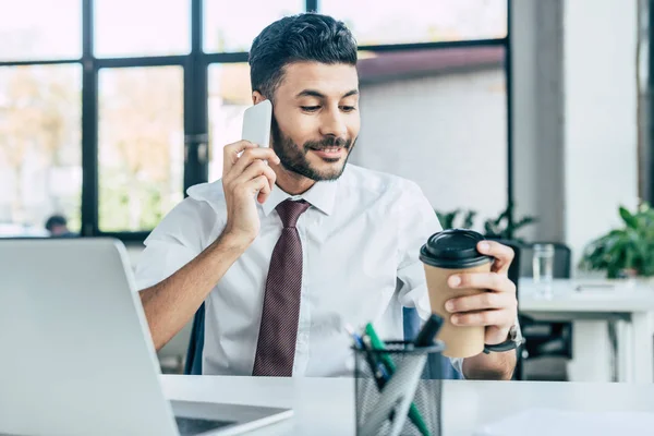 Sonriente hombre de negocios sosteniendo café para llevar mientras habla en el teléfono inteligente - foto de stock