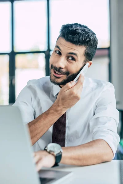Selective focus of cheerful businessman using laptop while talking on smartphone — Stock Photo
