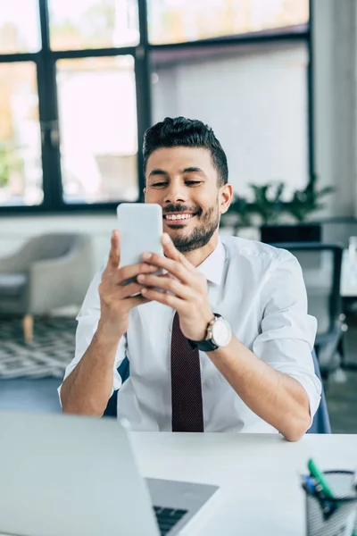 Enfoque selectivo de hombre de negocios alegre utilizando el teléfono inteligente mientras está sentado en el lugar de trabajo - foto de stock