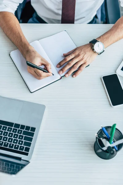 Partial view of businessman writing in notebook at workplace near laptop and smartphone — Stock Photo