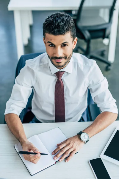 Schöner Geschäftsmann lächelt in die Kamera, während er am Arbeitsplatz in Notizbuch schreibt — Stockfoto