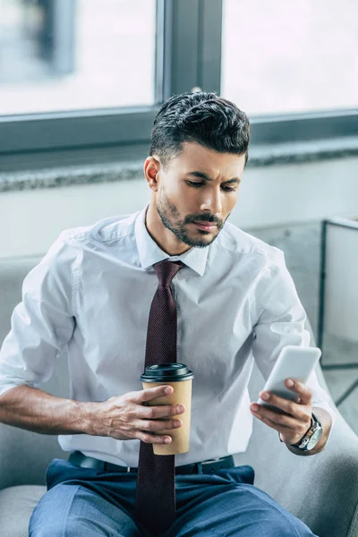 Hombre de negocios serio sentado en el sillón, el uso de teléfono inteligente y la celebración de café para ir - foto de stock